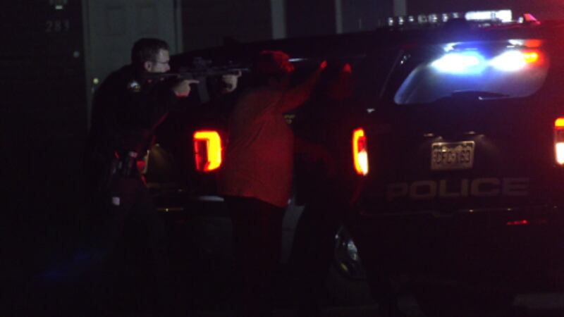 Armed GJ police officer and two other people stand behind police vehicle.